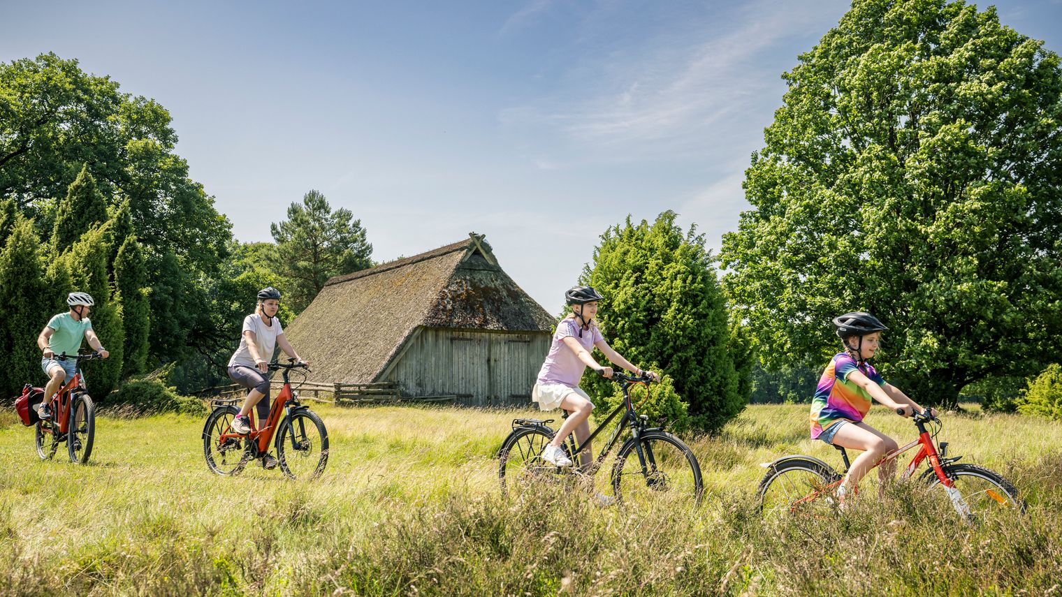 Familie mit zwei Erwachsenen und zwei Kindern fährt mit Fahrrädern durch das Naturschutzgebiet bei Bispingen, im Hintergrund ein Reetdachhaus und grüne Bäume